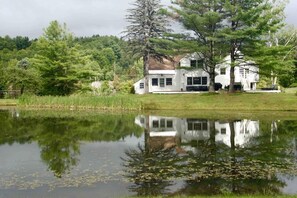 A white house with a large front porch, surrounded by trees and a pond.