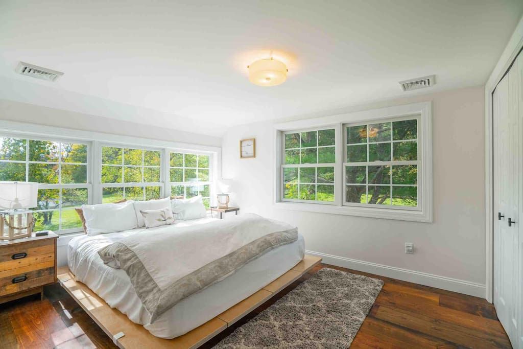 Un dormitorio con una cama grande, dos ventanas, una mesita de noche y piso de madera.