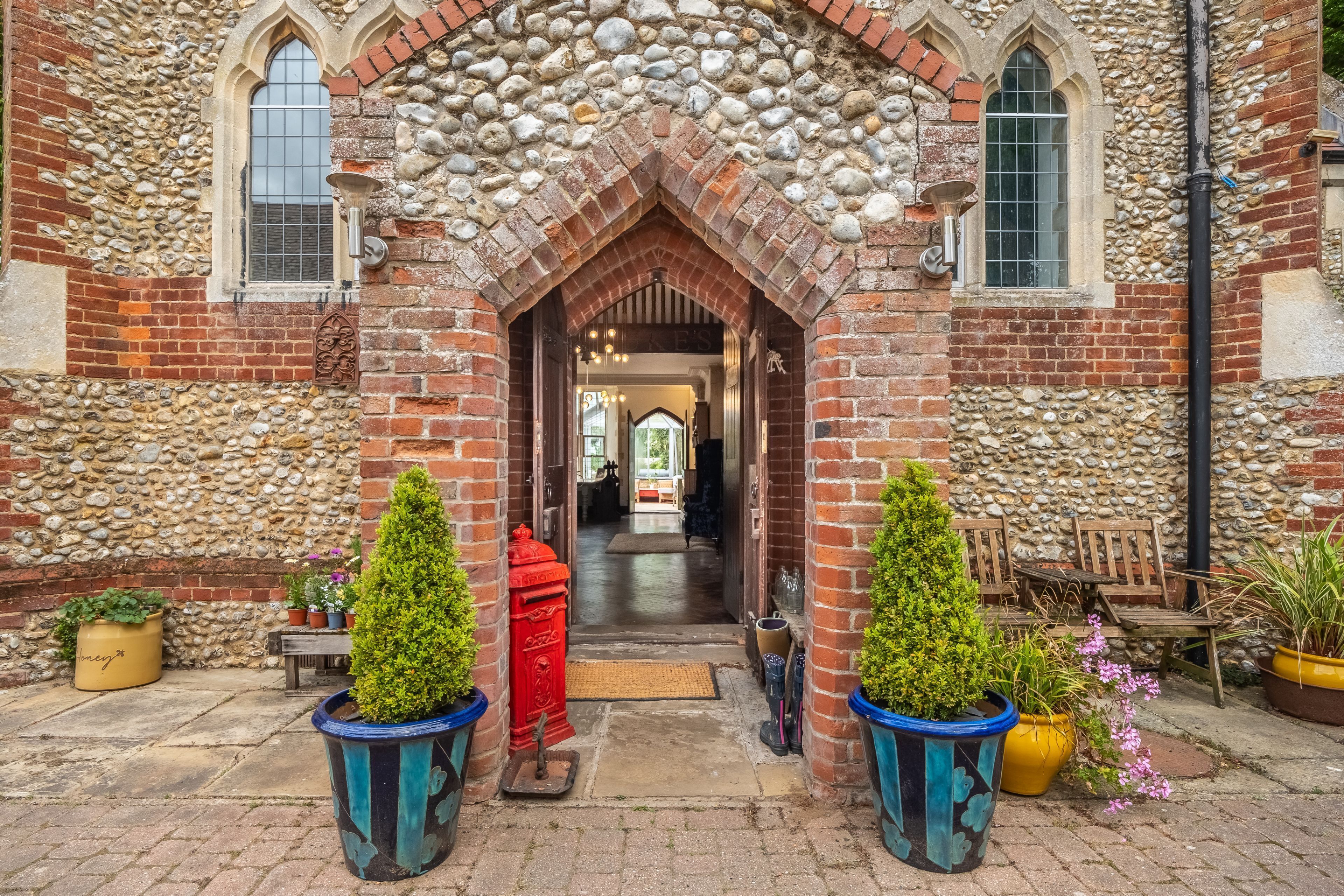 Un bâtiment en pierre avec une arche en briques qui mène à un couloir. Des plantes en pot et du mobilier d’extérieur sont disposés le long du chemin.