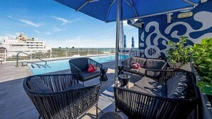 Zona de piscina en la azotea con sillones y una sombrilla, con vistas a un paisaje urbano.