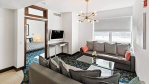 Una sala moderna con un sofá, una mesa de centro y un televisor. Se aprecia una alfombra estampada, un candelabro y una ventana con persianas.