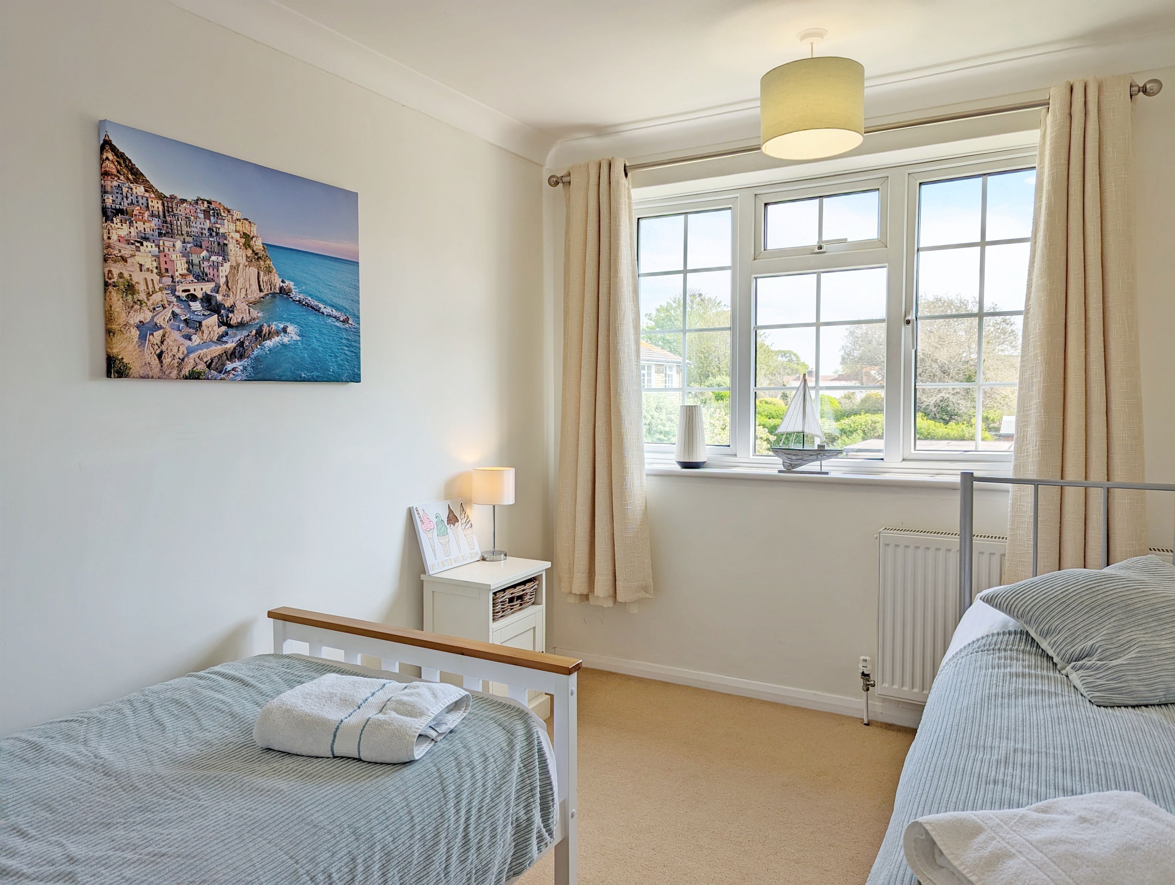 A bedroom with a bed, a bedside table with a lamp, a window with curtains, and a painting of a coastal scene.