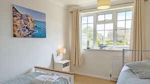 A bedroom with a bed, a bedside table with a lamp, a window with curtains, and a painting of a coastal scene.