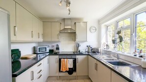 A modern kitchen with white cabinets, a microwave, a kettle, and a sink. There is a clock on the wall and a window with a view of trees.