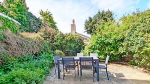 An outdoor patio with a stone table and chairs, surrounded by dense greenery and a brick chimney in the background.