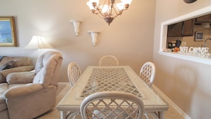 A dining area with a glass-top table, white chairs, and a chandelier. - CAPTAIN'S SUITE  Sea Coast (New Smyrna Beach)