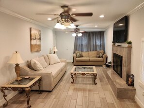 Una sala de estar con un sofá color beige, una mesa de centro de vidrio, chimenea y ventilador de techo.