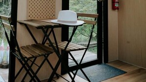 Una habitación con una mesa de madera, un sombrero blanco, una silla plegable negra, un extintor y vistas a árboles a través de un ventanal grande.