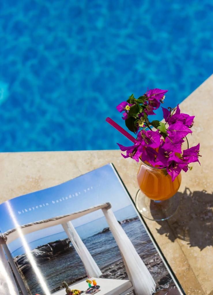 Un'area a bordo piscina con una piscina blu cristallina, un tavolo in vetro con una rivista e un vaso con fiori viola.