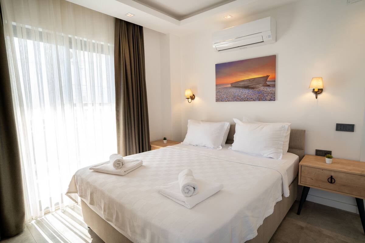 A hotel room with a large bed, two bedside tables, an air conditioner, a wall-mounted lamp, and a painting of a sunset over a beach.