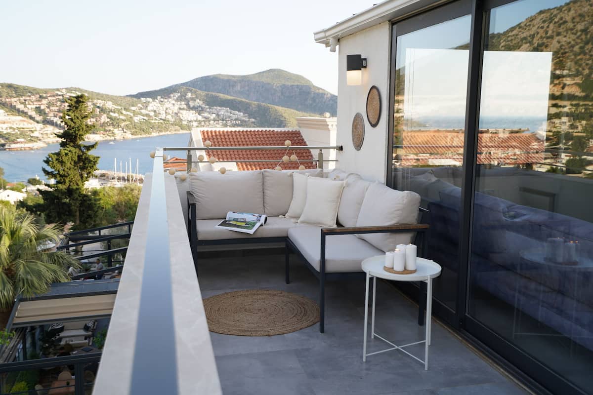A balcony with a sofa, a small table, and a view of the sea and mountains.