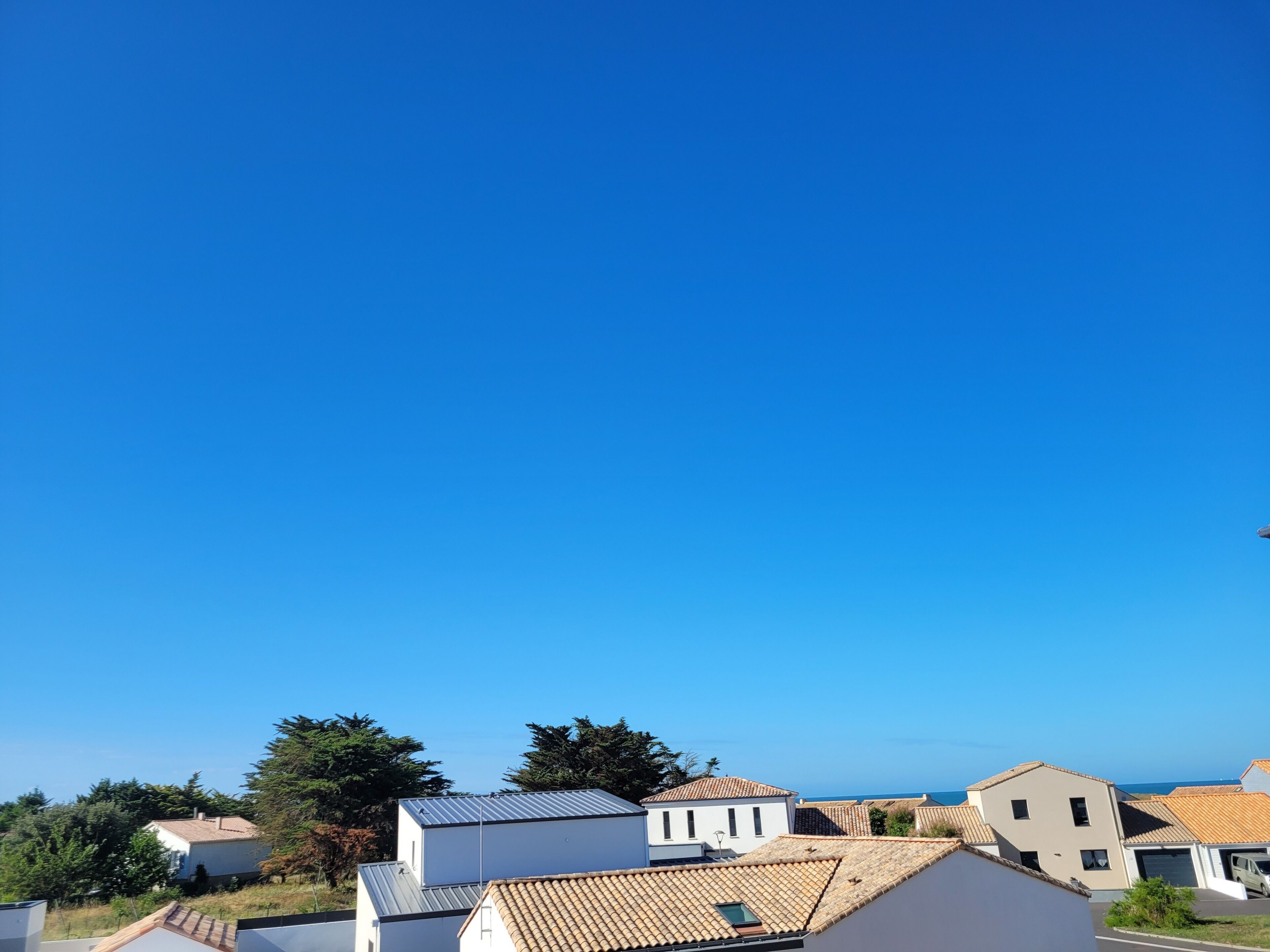 Zona residencial con casas de diversos estilos de tejado, un cielo azul despejado y vista al mar al fondo.