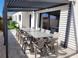 Un espacio de comedor al aire libre techado con una mesa larga y sillas, una pérgola en lo alto y vista a una calle residencial.