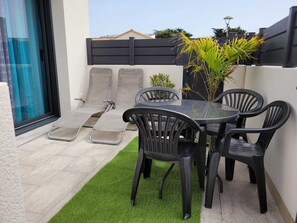 Un patio con puerta de vidrio, una mesa pequeña con sillas y una zona de césped artificial verde.