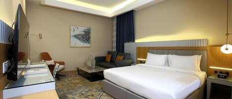 A hotel room with a large bed, a desk with a computer, a sofa, and a TV.
