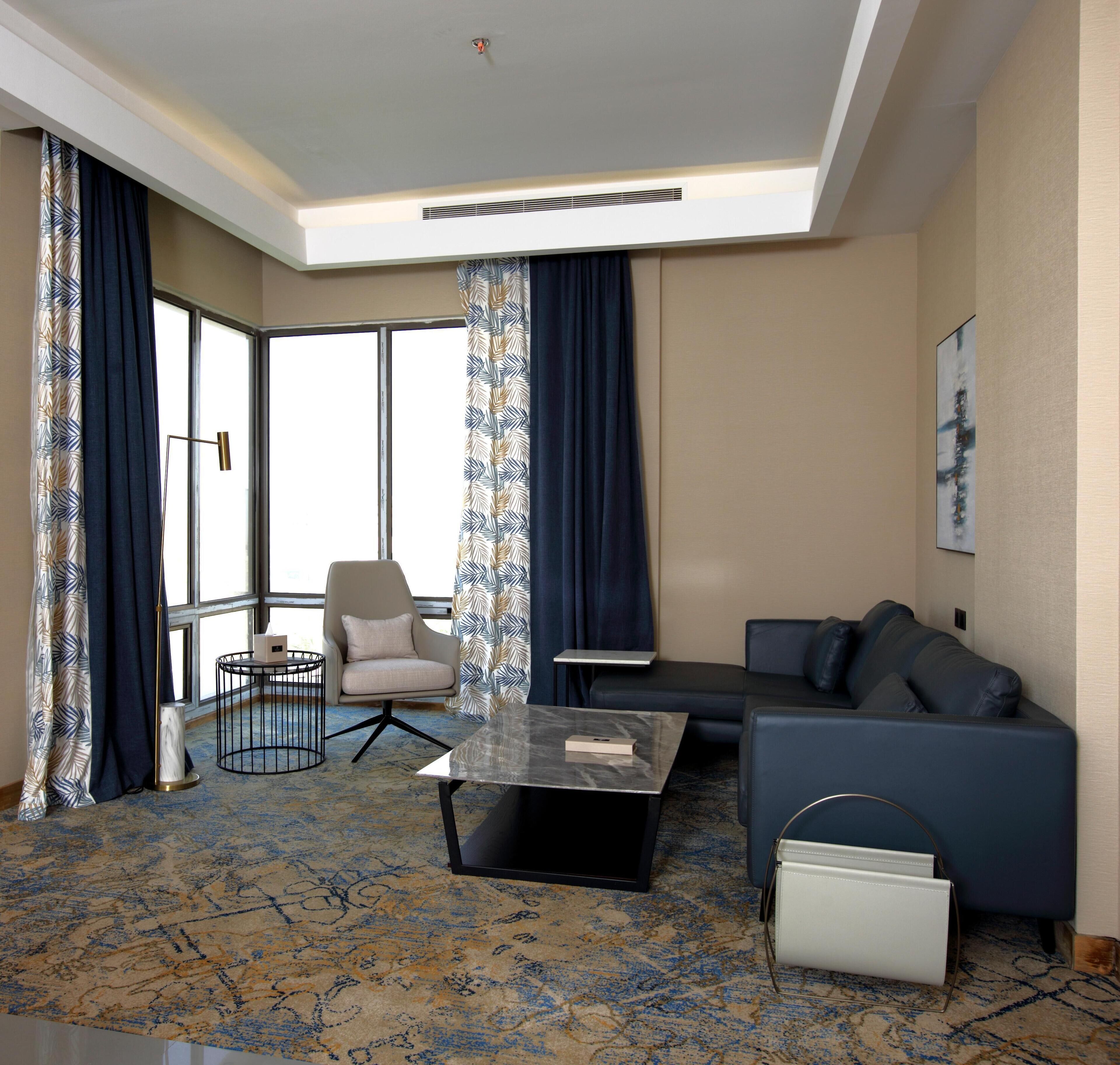 A modern hotel room with a sofa, armchair, and a small table. There is a large window with curtains, a ceiling light, and a wall-mounted air conditioning unit.