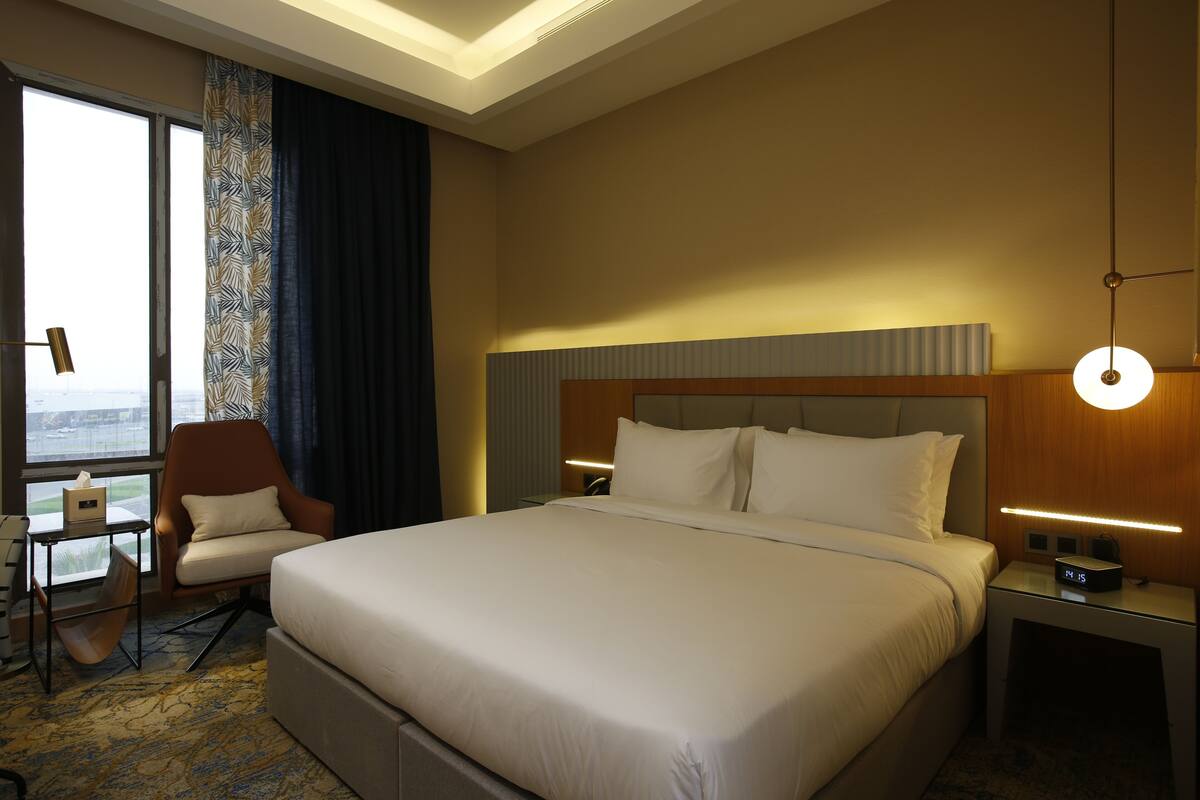 A hotel room with a large bed, a chair, a desk, and a nightstand with a lamp.