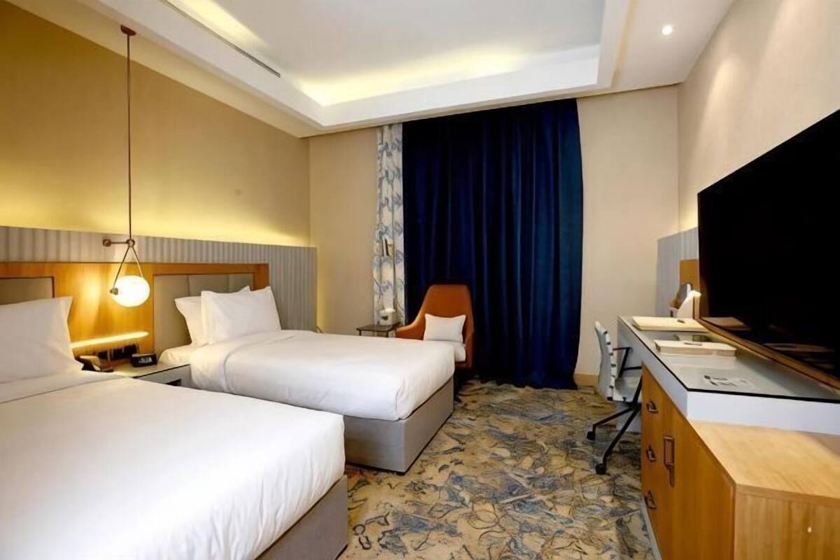 A hotel room with two beds, a desk, a TV, and a large window with curtains.