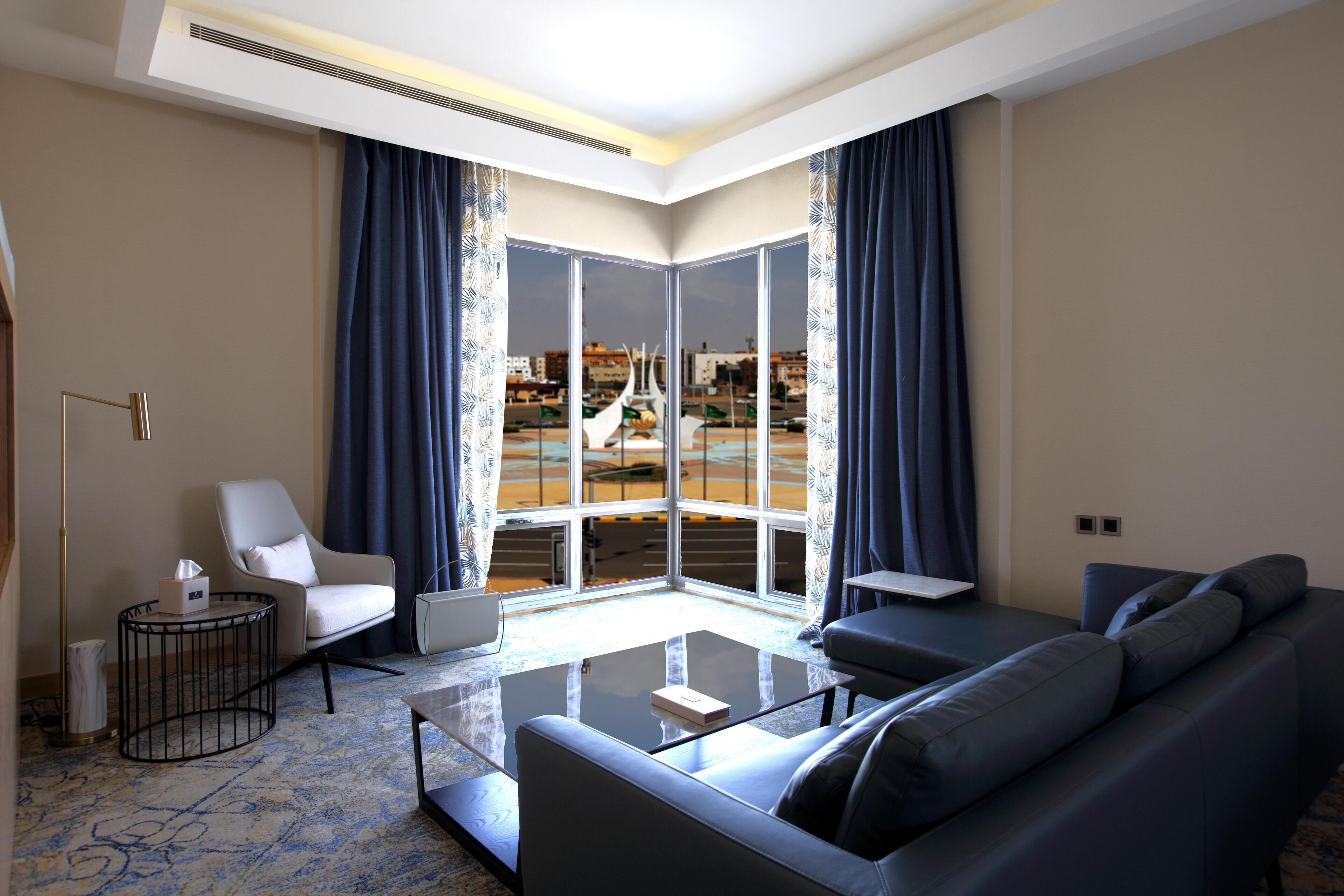 A modern living room with a glass coffee table, a sofa, a chair, and a view of a cityscape through large windows.