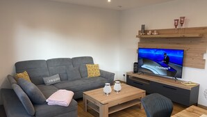 A modern living room with a grey sofa, a wooden coffee table, a TV on a wooden stand, and a small bookshelf.