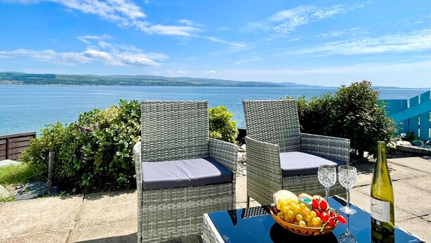 A table with a bottle of wine, a bowl of fruit, and two glasses, set against a scenic view of a body of water and hills. - Arthur's Seat,  elevated cottage, sea views (Dunoon)