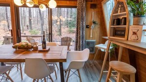 A wooden cabin interior with a dining table, chairs, a chandelier, and a view of the forest through large windows. - Lake Naomi A-Frame Cabin, Pet Friendly, (Pocono Pines)