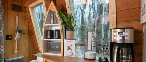 L’intérieur d’un chalet en bois comprend un coin cuisine équipé d’une machine à café, d’une cafetière Chemex et d’une carafe en verre.