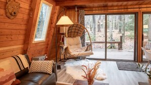 A cozy cabin interior with a wooden interior, a cushioned chair, a sofa, and a large window offering a forest view. - Lake Naomi A-Frame Cabin, Pet Friendly, (Pocono Pines)