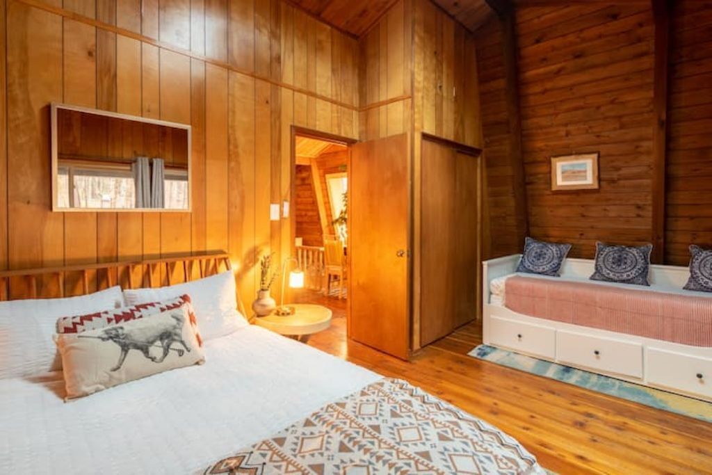 Camera da letto in una baita di legno, con un letto, una panca con cuscini, uno specchio e una porta che dà su una scala.