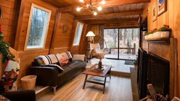 A cozy cabin interior with a wooden ceiling, a fireplace, a sofa, a lamp, and a view of the forest through large windows. - Lake Naomi A-Frame Cabin, Pet Friendly, (Pocono Pines)