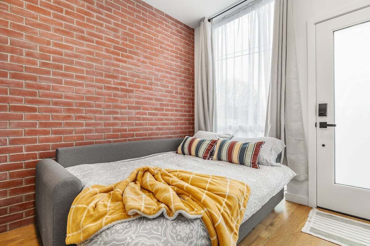 Een moderne slaapkamer met een bakstenen muur, een grijze bank, een bed met beddengoed met een patroon, en een raam met dunne gordijnen.