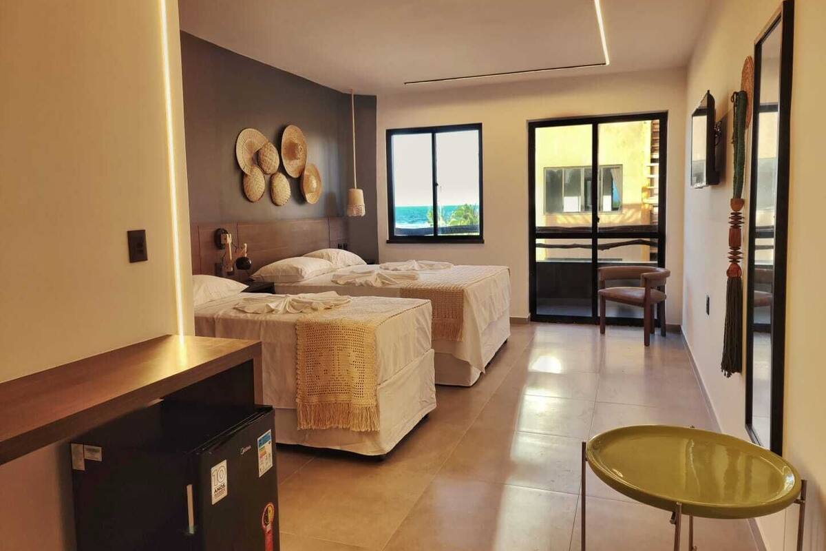 Hotelkamer met twee bedden, een kleine tafel, een stoel, een groot raam met uitzicht op zee en een balkon met schuifdeur.