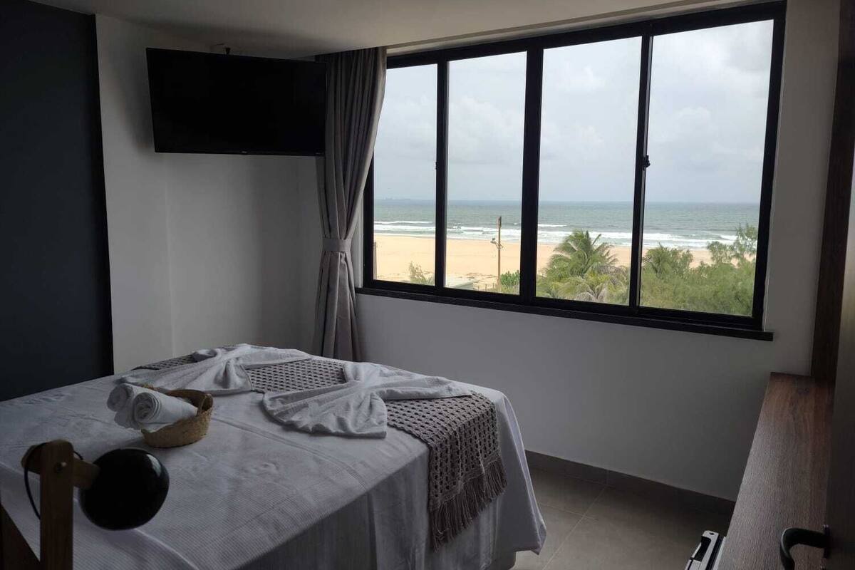 Een slaapkamer met een groot raam met uitzicht op het strand, een bed met handdoeken en een flatscreen televisie aan de muur.