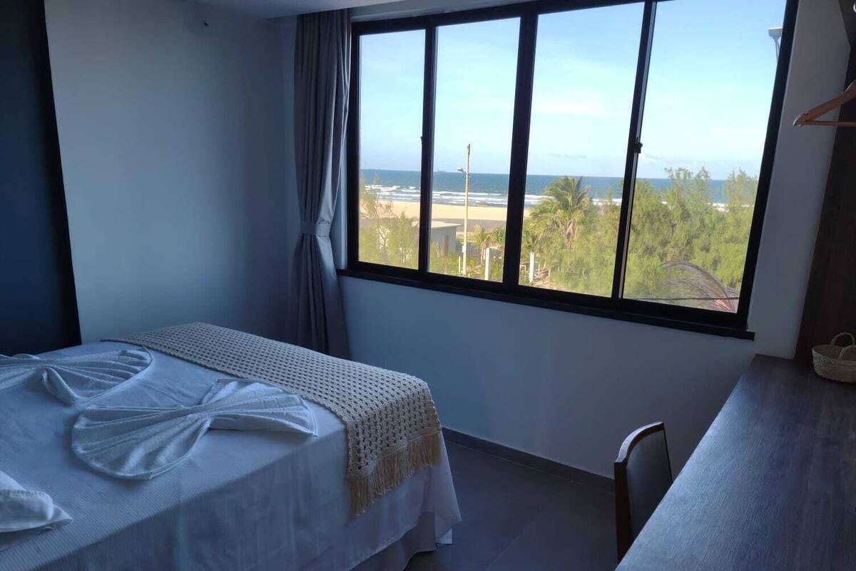 Een hotelkamer met een bed, een bureau en een groot raam met uitzicht op het strand en de oceaan.