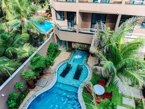 A swimming pool with two lounge chairs, surrounded by palm trees and a building with large windows. - Maly Boutique Hotel (Fortaleza)