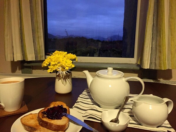 A breakfast setup with a teapot, cups, a small vase with yellow flowers, and a plate with toast and jam.