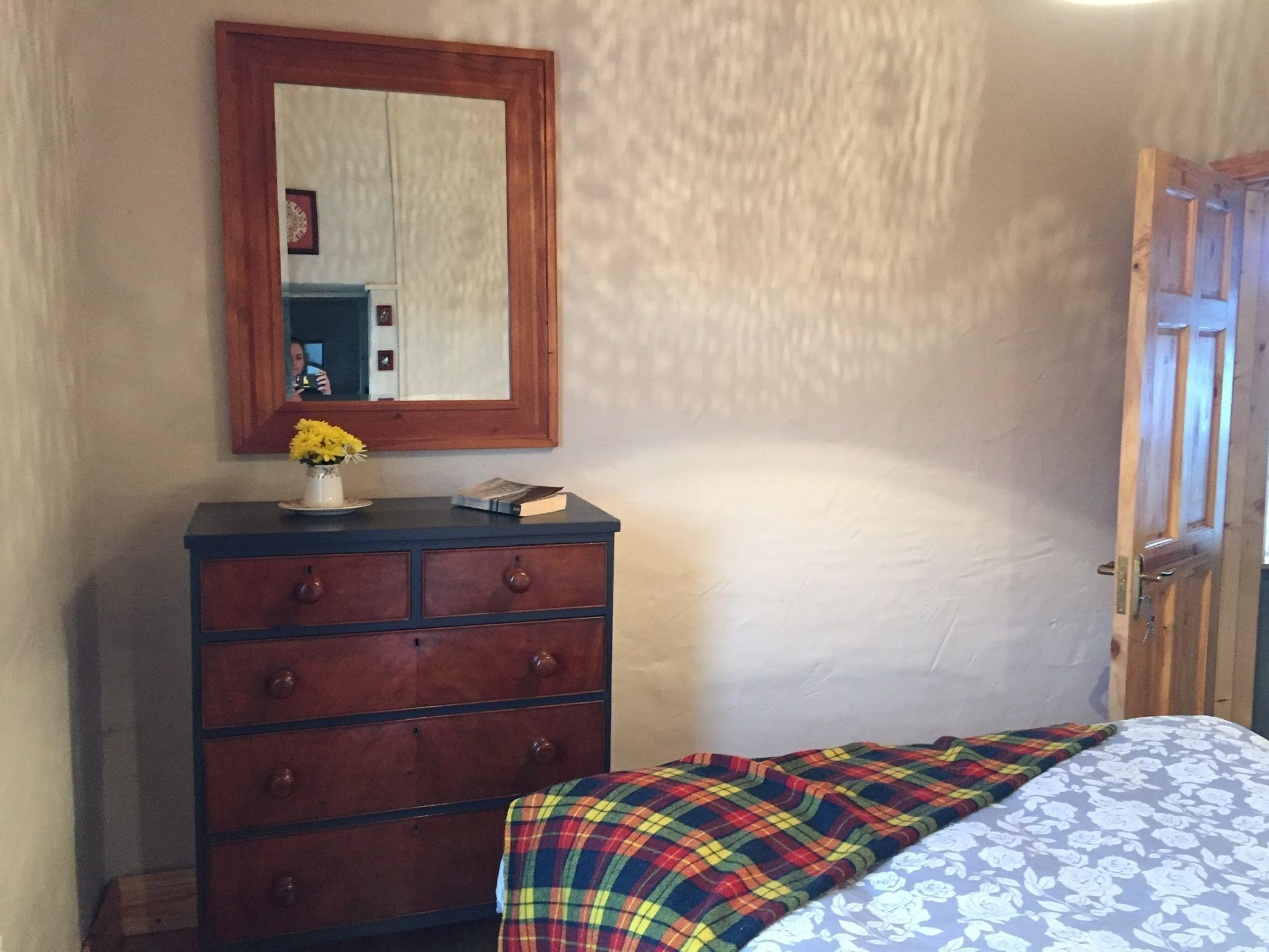 Een slaapkamer met een houten dressoir, een spiegel, een bed met een bloemenprint en een deur.