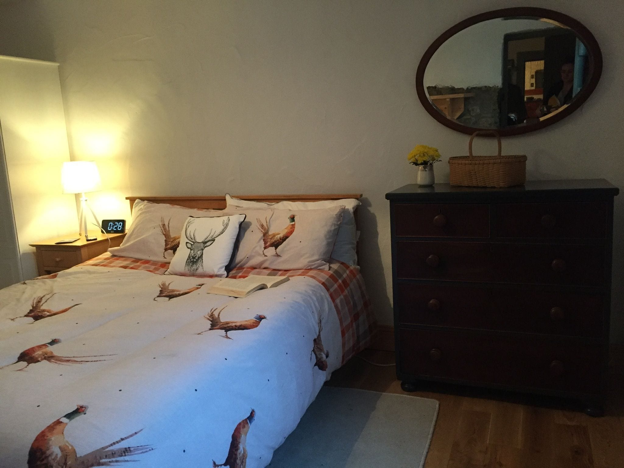 A bedroom with a bed featuring a deer and rooster print, a bedside lamp, a dresser, and a mirror.