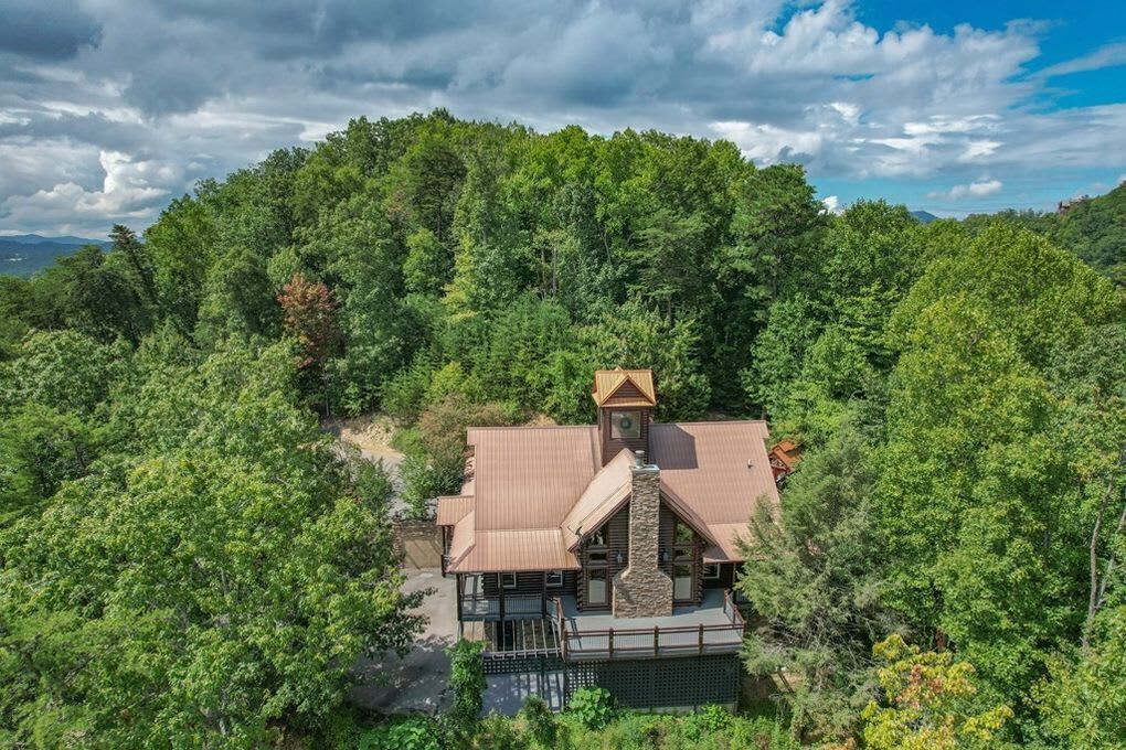 A large, rustic cabin surrounded by dense forest, with a prominent chimney and a small tower-like structure.