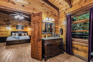 A wooden cabin interior with a bed, a dresser, a mirror, and a window with curtains.