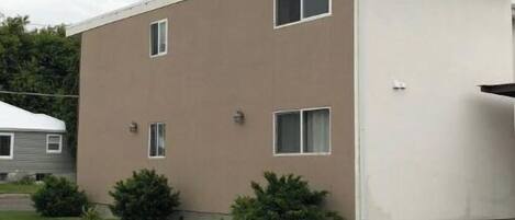 A two-story apartment building with a flat roof, a small front yard with bushes, and a visible side entrance.