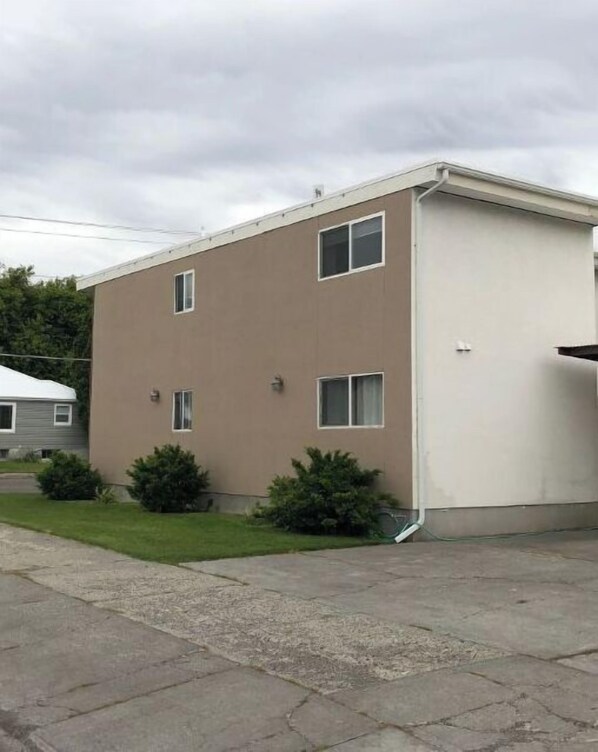 Un edificio de apartamentos de dos pisos con techo plano, un pequeño jardín delantero con arbustos y una entrada lateral visible.