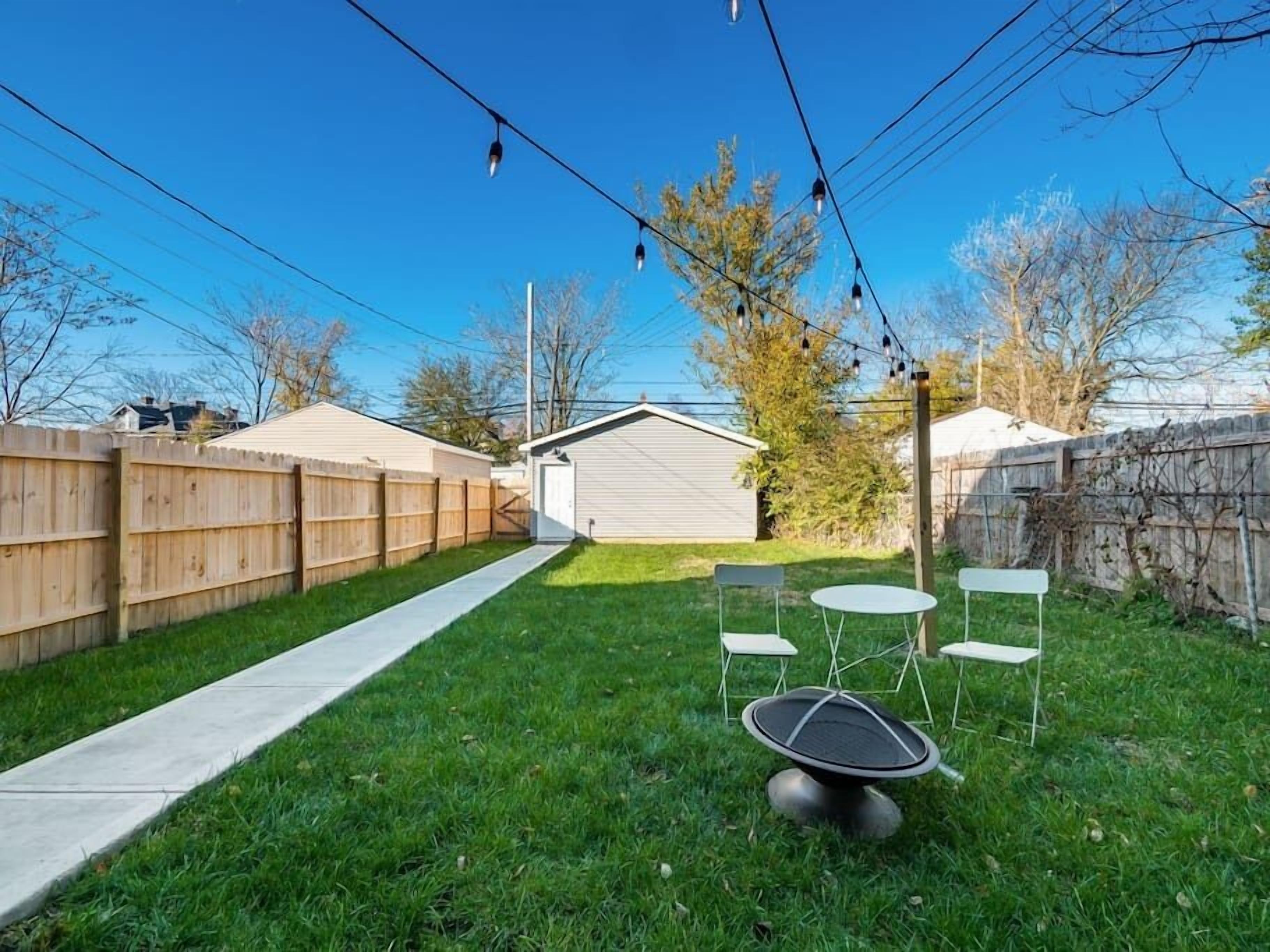 A backyard with a concrete pathway, a small shed, and a trampoline surrounded by white chairs.