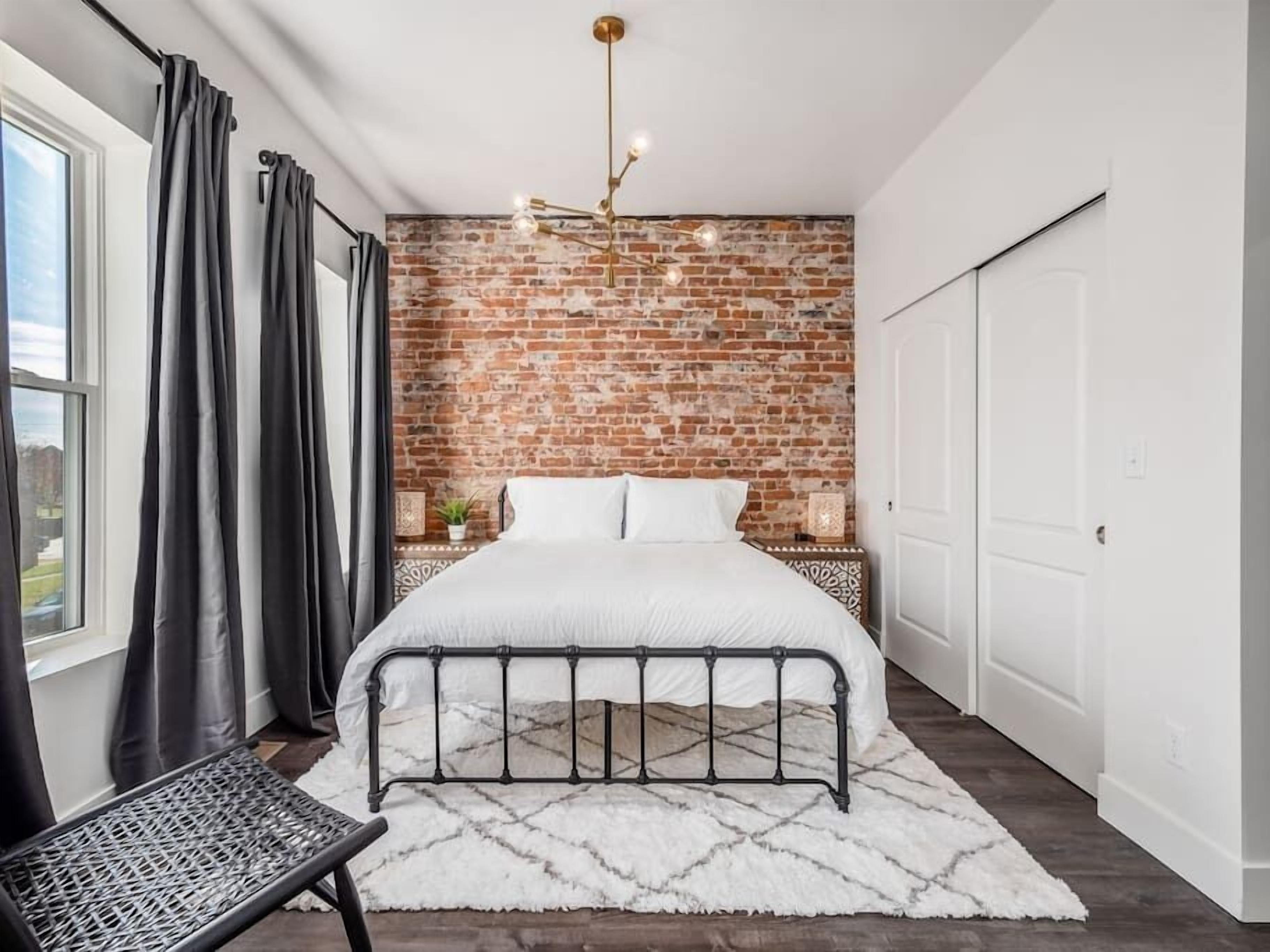 Un dormitorio con pared de ladrillo, cama con estructura metálica, alfombra blanca, un banco y una lámpara colgante.