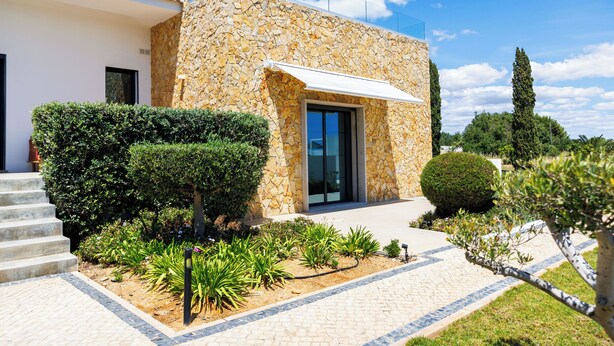 Ein modernes Haus mit Naturstein-Fassade, einer Laube und einem gepflegten Garten mit verschiedenen Pflanzen und einem gepflasterten Weg.