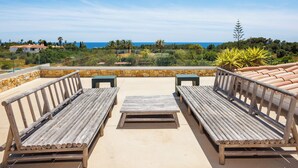 Eine Terrasse mit Holzbänken und -tischen, die auf eine malerische Landschaft mit Blick auf das Meer und entfernte Gebäude blickt.