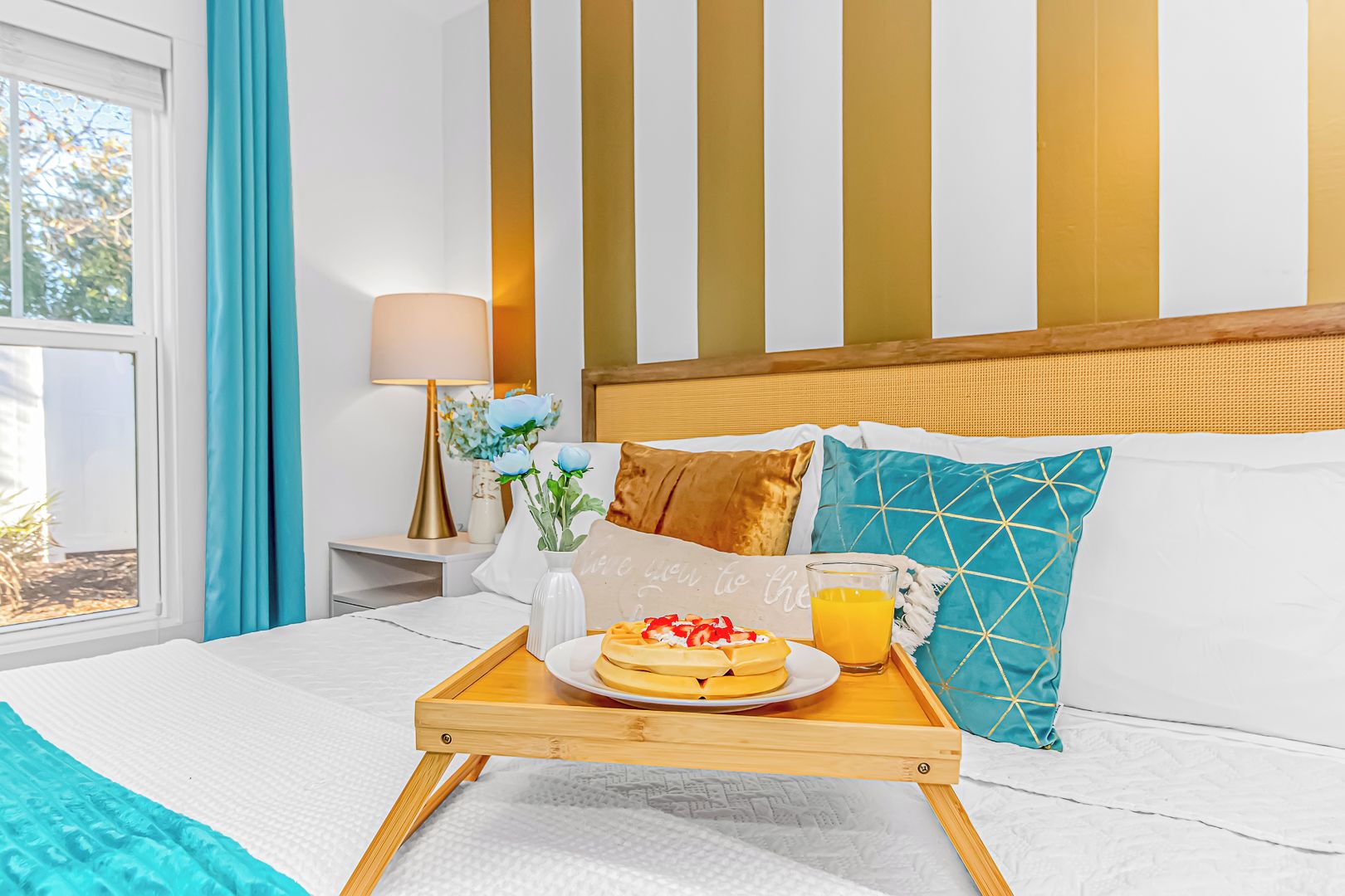 Quarto de hotel com cama, mesa de cabeceira com abajur, bandeja com itens de café da manhã e janela com cortinas.
