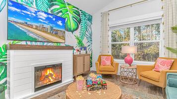 Una sala de estar con chimenea, un televisor montado en la pared, un sofá, una silla, una mesita con lámpara y una ventana con cortinas.