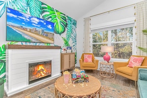 A living room with a fireplace, a television mounted on the wall, a sofa, a chair, a small table with a lamp, and a window with curtains.