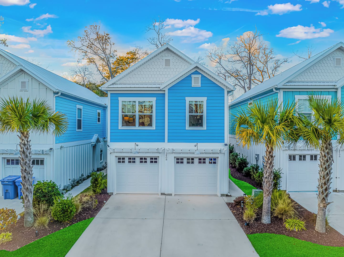 A residential area with three houses, each with a garage and a front yard with palm trees.